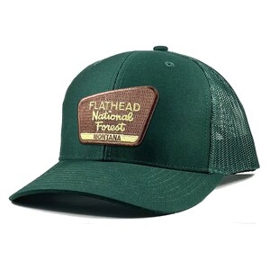 May include: A green trucker hat with a brown patch that says "Flathead National Forest Montana" in yellow lettering.