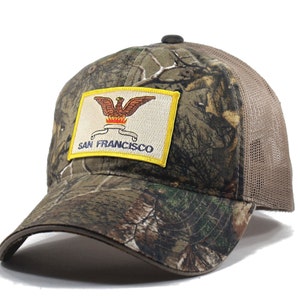 May include: A camouflage trucker hat with a yellow patch featuring the San Francisco city seal. The seal depicts a golden eagle with a red flame and a banner that reads "San Francisco".