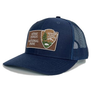 May include: A navy blue trucker hat with a brown patch featuring the Kenai Fjords National Park logo. The logo includes the text "Kenai Fjords National Park" and "National Park Service".