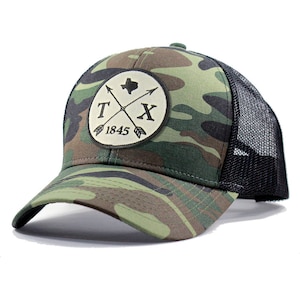 May include: A camouflage trucker hat with a black mesh back. The front of the hat features a round patch with a Texas state outline, crossed arrows, and the text "TX 1845".