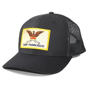 May include: Black trucker hat with a black mesh back. The front of the hat features a yellow patch with the San Francisco city seal, which includes a golden eagle with a red crown and a blue banner that reads "SAN FRANCISCO".