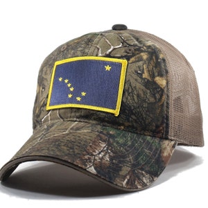 Peut inclure: Un chapeau de camionneur camouflage brun et vert avec un filet arrière brun. Le chapeau présente un patch brodé jaune du drapeau de l'État d'Alaska.