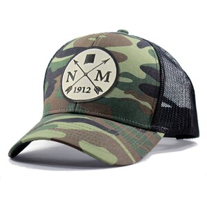 May include: A camouflage trucker hat with a black mesh back. The front of the hat features a round patch with a state outline, crossed arrows, and the text "NM 1912".