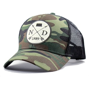 May include: A camouflage trucker hat with a black mesh back. The front of the hat features a circular patch with the letters "ND" and a crossed arrow design. The text "1889" is below the letters.