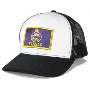May include: White trucker hat with a black mesh back. The front of the hat features a patch with the state seal of Kansas and the word "Kansas" in gold lettering.