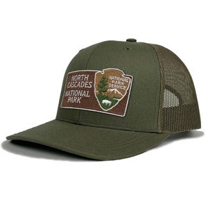 May include: Olive green trucker hat with a curved brim and mesh back. The front features a rectangular patch with the text "North Cascades National Park" and the National Park Service logo, including a tree, mountain, and bear.