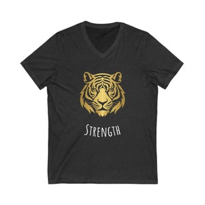 Puede incluir: Una camiseta negra de cuello en V con un gráfico de cabeza de tigre dorado y la palabra "STRENGTH" impresa en blanco debajo del gráfico.