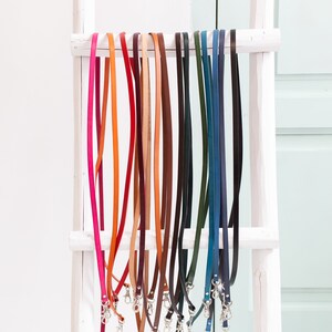 K&ouml;nnte beinhalten: Eine Sammlung von Lederb&auml;ndern in verschiedenen Farben, darunter Rot, Orange, Gelb, Gr&uuml;n, Blau und Braun, die an einer wei&szlig;en Leiter h&auml;ngen.