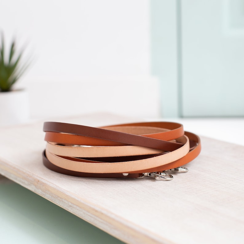 K&ouml;nnte beinhalten: Ein Stapel aus vier Lederriemen in den Farben Braun, Orange und Beige. Die Riemen sind kreisf&ouml;rmig angeordnet und haben silberne Metallschlie&szlig;en.