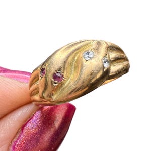 May include: A gold ring with a snake design. The ring features two clear gemstones and two red gemstones. The ring is held by a person with pink nail polish.