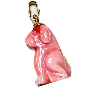 May include: A carved pink coral pendant in the shape of a seated dog, featuring a smooth, polished finish with natural colour variations. The pendant is attached to a simple gold-toned metal bail at the top, designed for use on a necklace chain.