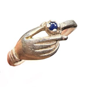 May include: A silver-toned ring featuring a hand design, with the index finger pointing towards a round, dark blue gemstone. The ring has a detailed, textured appearance, and the gemstone is set in a raised bezel.