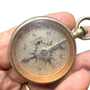May include: A vintage, round compass with a silver-colored metal casing. The compass face has directional markings, degree indicators, and the text "LEEDAWL." The needle points towards the southeast. A small loop is attached to the top for carrying.