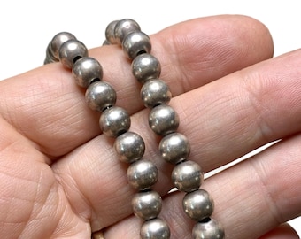May include: Close-up of a silver beaded necklace. The necklace features multiple strands of round, metallic beads. The beads are a muted silver colour and appear to be strung together with a dark cord. The necklace is held in a person's hand.