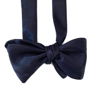 May include: A navy blue bow tie made from a shiny, satin-like fabric. The bow is tied, with two long straps hanging down. The background is white.
