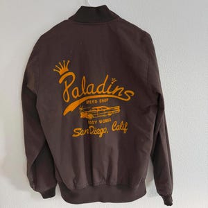 May include: A brown bomber jacket with a zip-up front and a dark brown collar and cuffs. The back of the jacket features the words "Paladins Speed Shop Body Works San Diego, Calif" in orange script and a car graphic.