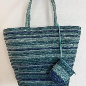 May include: A woven tote bag and matching small pouch in shades of blue and green. The bag has a rectangular shape with a wide opening and sturdy handles. The pouch is attached with a thin cord. The bag is made of a woven material.