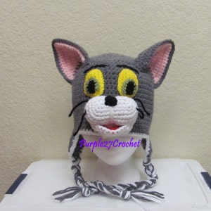 May include: Gray crocheted Tom and Jerry character hat with yellow eyes, white nose, and a pink nose. The hat has a long gray and white braided cord.