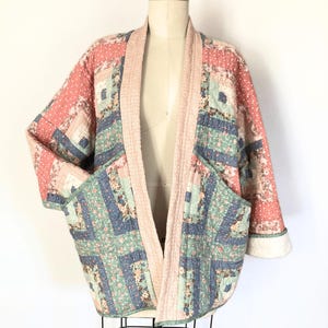 May include: A quilted jacket in shades of pink, blue, and green, displayed on a mannequin. The jacket features a patchwork design with floral and geometric patterns, a light pink collar, and two front pockets. The sleeves have a white cuff.