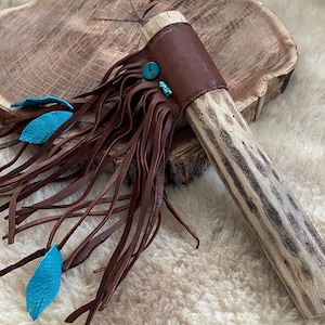 Puede incluir: Una pipa ceremonial artesanal con un tallo de madera clara con rayas marrones oscuras. La pipa tiene una envoltura de cuero marrón con detalles turquesa y largas flecos de cuero marrón, y dos plumas de cuero turquesa.