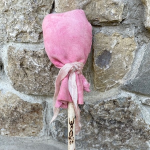 Könnte beinhalten: Ein handgefertigter Holzstab mit einem rosafarbenen Wildleder-Kopf und einem dekorativen Design. Der Stab ist mit einem rosa Stoffband und einer geschnitzten Figur verziert. Das Holz hat ein natürliches, gealtertes Aussehen.