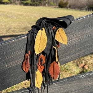 May include: A black leather fringe accessory featuring multiple dangling leather strips adorned with several small, flat, leaf-shaped cutouts in shades of mustard yellow and burnt orange, each with a central etched line detail.