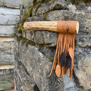 Puede incluir: Una pipa de madera hecha a mano con una banda de cuero con un diseño de plumas. La pipa tiene una franja de tiras de cuero y plumas decorativas en tonos marrones y negros. La pipa está apoyada en un muro de piedra.