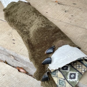 May include: A rolled-up, olive-green sheepskin with a white interior and a patterned fabric lining. The sheepskin is secured with three black toggles. The fabric lining features a geometric design in shades of green, beige, and black.