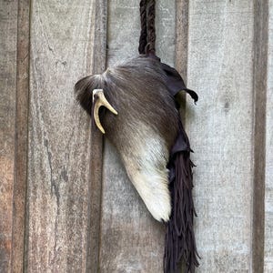 May include: A rustic wall hanging made from natural hide, with a cream and brown colour palette. A small antler is attached to the hide. It is suspended by a braided leather strap and has a dark brown fringe.