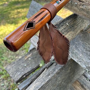 May include: A wooden flute with a polished brown finish, adorned with turquoise inlays and a leather binding. Decorative cutouts and two brown leather feathers enhance its design. The flute rests on weathered wooden planks, adding to its rustic aesthetic.