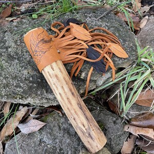 May include: A handcrafted wooden pipe with a tan leather wrap featuring a feather design. The pipe has a natural wood finish and is adorned with tan leather fringe and black accents. The pipe is resting on a stone.