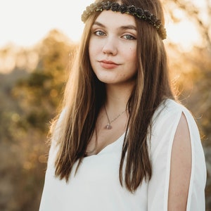 May include: A woman with long brown hair wearing a black rose crown and a white top with open sleeves. She is also wearing a necklace with a pendant. The background is blurred with warm, golden tones.