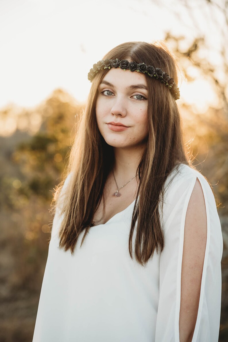 May include: A woman with long brown hair wearing a black rose crown and a white top with open sleeves. She is also wearing a necklace with a pendant. The background is blurred with warm, golden tones.