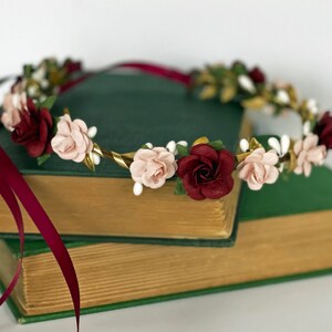 Op de afbeelding: Een bloemenkrans met afwisselend bordeauxrode en lichtroze rozen, afgewisseld met kleine witte knopjes en goudkleurige blaadjes. Het ronde hoofddeksel is versierd met lange, vloeiende bordeauxrode satijnen linten aan de zijkanten.