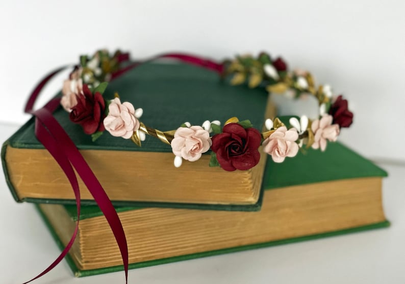 Op de afbeelding: Een bloemenkrans met afwisselend bordeauxrode en lichtroze rozen, afgewisseld met kleine witte knopjes en goudkleurige blaadjes. Het ronde hoofddeksel is versierd met lange, vloeiende bordeauxrode satijnen linten aan de zijkanten.