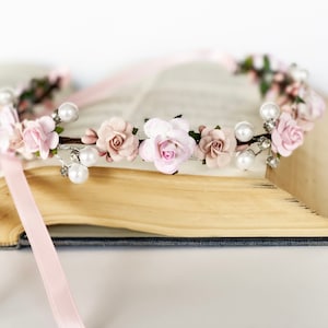 May include: A floral crown featuring delicate pink paper roses, pearls, and sparkling accents. The crown is adorned with a light pink ribbon and rests on an open book. This headpiece is perfect for weddings, festivals, or special occasions.