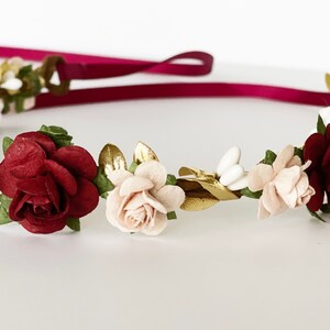 Op de afbeelding: Een bloemenkrans met kunstmatige bordeauxrode en zachtroze rozen, afgewisseld met kleine witte knopjes en metallic gouden blaadjes. Het geheel is bevestigd aan een lang, donkerbordeauxrood lint dat aan de achterkant van de krans uitsteekt.
