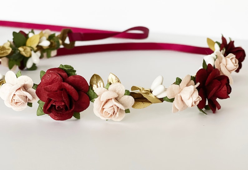 Op de afbeelding: Een bloemenkrans met kunstmatige bordeauxrode en zachtroze rozen, afgewisseld met kleine witte knopjes en metallic gouden blaadjes. Het geheel is bevestigd aan een lang, donkerbordeauxrood lint dat aan de achterkant van de krans uitsteekt.