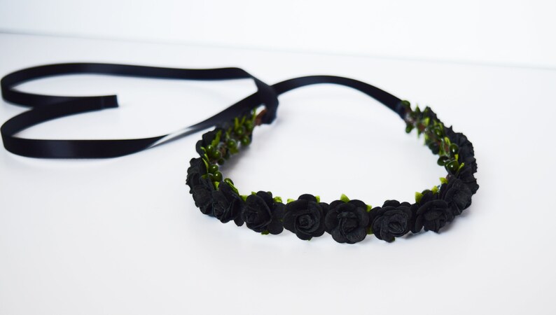 May include: A black floral crown with black roses and green leaves. The crown is made of fabric and has a black satin ribbon attached for tying. The crown is suitable for weddings, parties, or everyday wear.