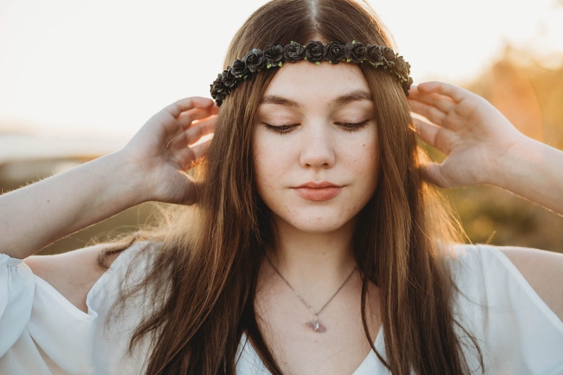 May include: A woman wearing a dark floral crown with closed eyes. The crown is made of small, dark roses. She is wearing a white top and a silver necklace with a pendant. The background is blurred with warm sunlight.