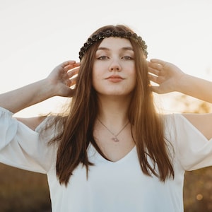 May include: A woman wearing a white blouse with a V-neck and long sleeves. She is wearing a black floral crown and a silver necklace. Her long brown hair frames her face as she holds her hands up near her head.