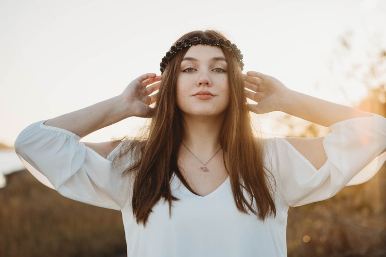 May include: A woman wearing a white blouse with a V-neck and long sleeves. She is wearing a black floral crown and a silver necklace. Her long brown hair frames her face as she holds her hands up near her head.