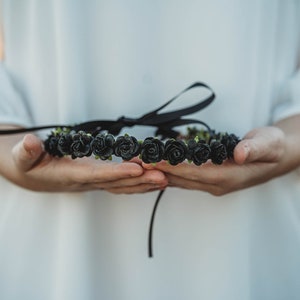 May include: A black rose flower crown with a black ribbon tie. The floral crown is held in open hands, with a white background. The roses are clustered together, creating a full, decorative headpiece.