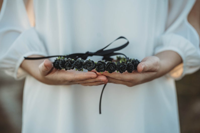 May include: A black rose flower crown with a black ribbon tie. The floral crown is held in open hands, with a white background. The roses are clustered together, creating a full, decorative headpiece.