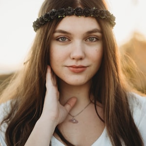 May include: A young person with long brown hair and a black floral crown. They are wearing a white top and a silver necklace with a pendant. The background is blurred with warm tones.