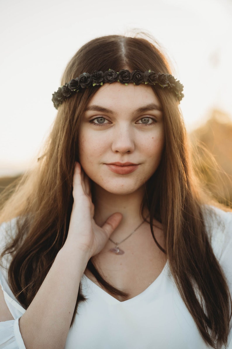 May include: A young person with long brown hair and a black floral crown. They are wearing a white top and a silver necklace with a pendant. The background is blurred with warm tones.
