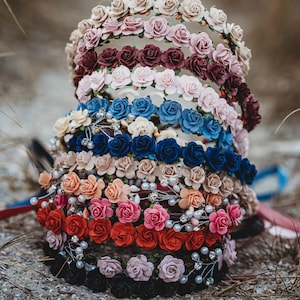 May include: A collection of floral headbands in various colors, including cream, pink, burgundy, blue, and red. Each headband is adorned with small fabric roses and some are accented with pearls. The headbands are arranged on a sandy surface.