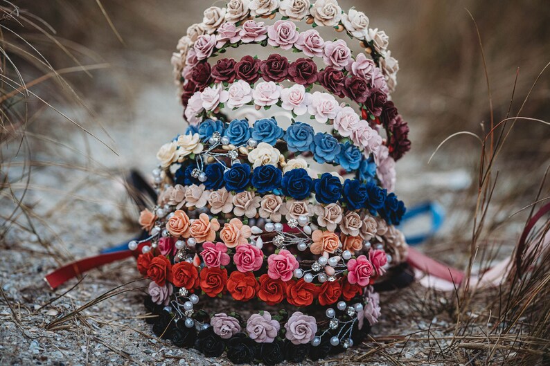May include: A collection of floral headbands in various colors, including cream, pink, burgundy, blue, and red. Each headband is adorned with small fabric roses and some are accented with pearls. The headbands are arranged on a sandy surface.