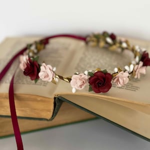 Op de afbeelding: Een bloemenkrans met afwisselend diepbordeauxrode en zachtroze rozen, geaccentueerd met kleine witte besjes en groene blaadjes op een goudkleurige draadbasis. Het accessoire is voorzien van lange, vloeiende bordeauxrode linten aan de uiteinden.