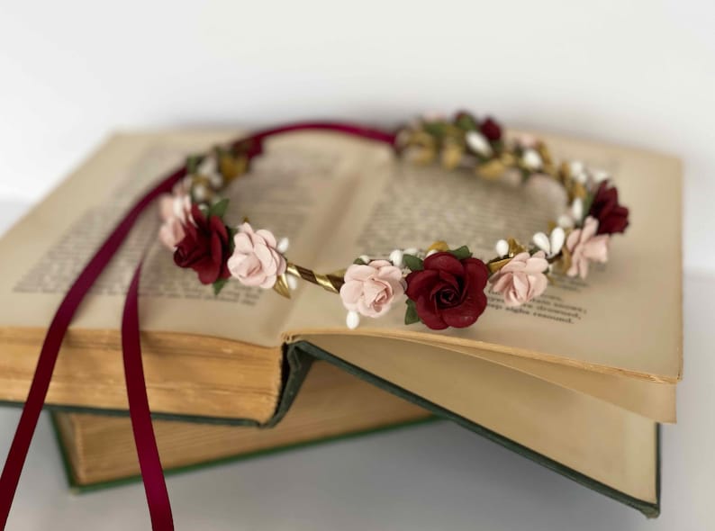 Op de afbeelding: Een bloemenkrans met afwisselend diepbordeauxrode en zachtroze rozen, geaccentueerd met kleine witte besjes en groene blaadjes op een goudkleurige draadbasis. Het accessoire is voorzien van lange, vloeiende bordeauxrode linten aan de uiteinden.